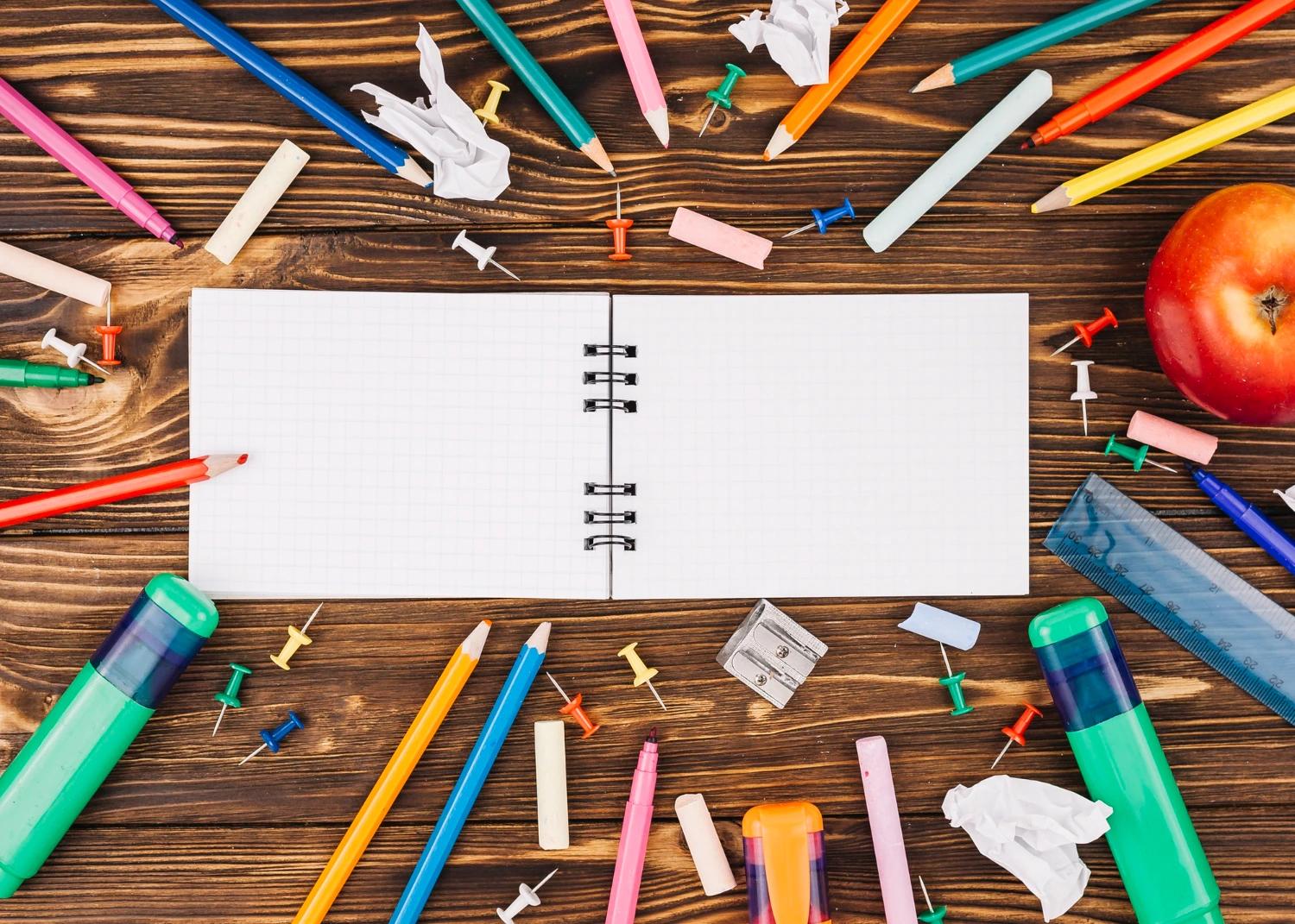 Uma mesa de madeira com um caderno aberto no centro, rodeado por diversos materiais escolares coloridos. Há lápis de várias cores, marcadores, apontadores, pinos de papel, borrachas e uma régua. Um lápis apontador e alguns papéis amassados também estão espalhados pela mesa. Ao lado direito, há uma maçã vermelha, simbolizando o ambiente de estudo ou planejamento educacional. O cenário sugere organização e preparação para atividades de aprendizado. Conceito: como criar um plano de aula.