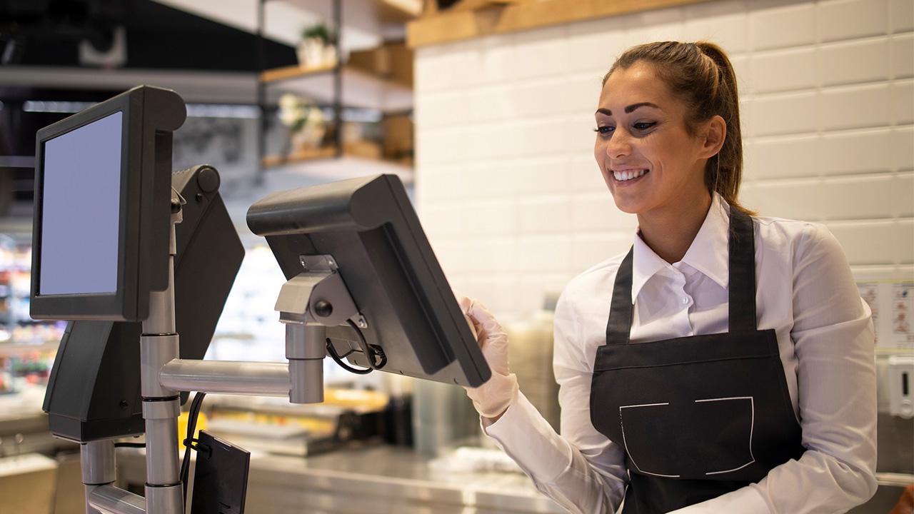 Uma mulher em frente a um caixa eletrônico pesando um produto em referência às competências de um operador de caixa.