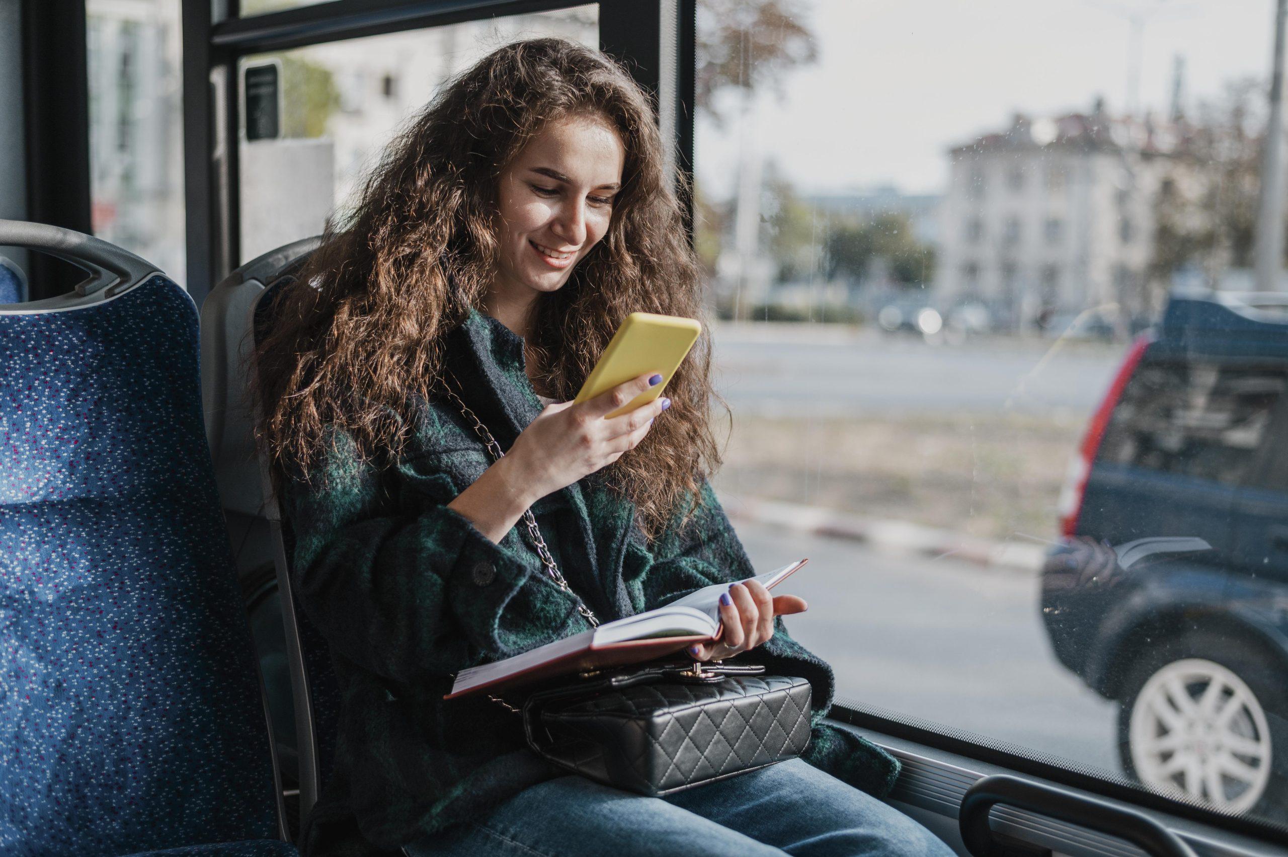 Jovem sorrindo enquanto estuda pelo celular dentro de um ônibus, com um caderno aberto no colo e vista urbana ao fundo.