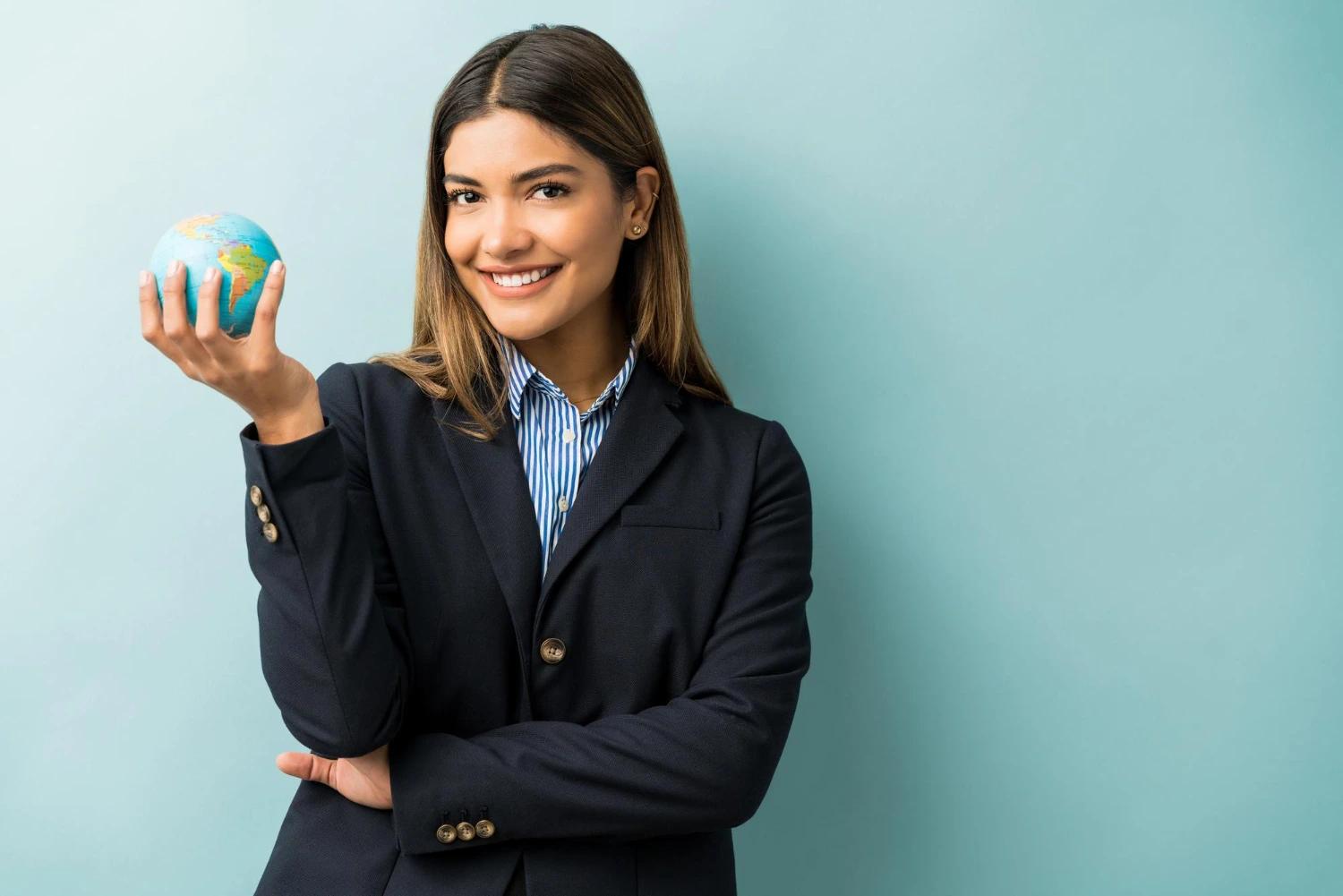 Uma jovem executiva de terno preto sorri segurando um pequeno globo terrestre na mão, em frente a um fundo azul claro. A cena transmite liderança e responsabilidade global, representando o papel dos profissionais especializados em ESG na construção de soluções sustentáveis para empresas e organizações.